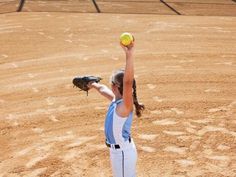 a woman in blue and white uniform throwing a ball on dirt field next to bleachers