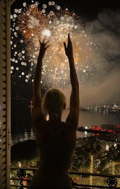 a woman standing in front of a window with her hands up to the sky as fireworks go off