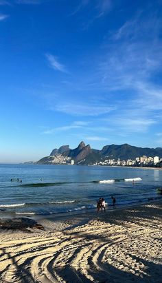 some people are walking on the beach by the water and mountains in the distance with blue skies