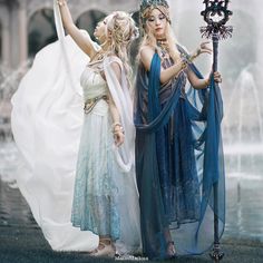 two beautiful women dressed in costume standing next to each other near a fountain with water spouting from it