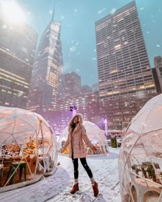 a woman standing in the snow between two clear domes with buildings in the back ground