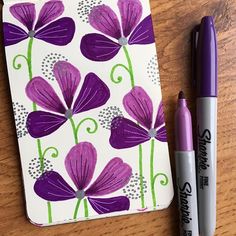 two pens sitting on top of a wooden table next to a notepad with purple flowers