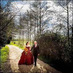 a man and woman dressed in red walking down a dirt road next to tall trees