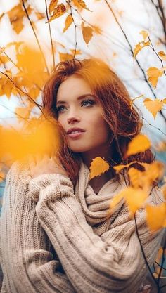 a woman with red hair is standing in front of yellow leaves and looking at the camera