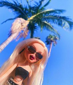 a barbie doll with blonde hair and sunglasses standing in front of a palm tree on a sunny day