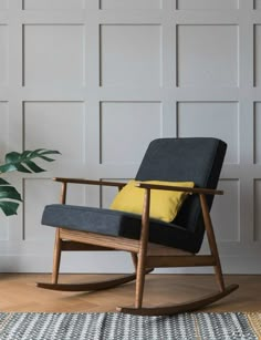 a grey rocking chair with yellow pillows in front of a white wall and wooden floor