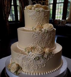 a three tiered wedding cake with white flowers on the top and sides, sitting on a table in front of a window