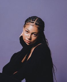 a woman with braids sitting in front of a purple wall and looking at the camera