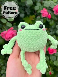 a hand holding a small green crocheted frog toy in front of pink flowers