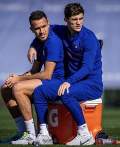 two men in blue uniforms sitting on an orange cooler with their arms around each other