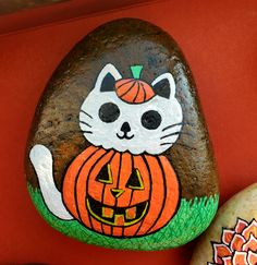 a rock with a cat painted on it next to an orange pumpkin and flower pot