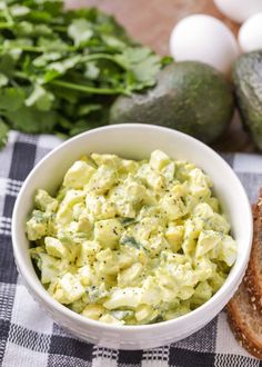 a white bowl filled with food next to bread and avocado on a checkered table cloth