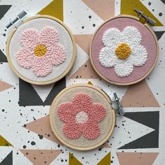 three hand embroidered flowers are sitting on top of a patterned tablecloth, one is pink and the other is white