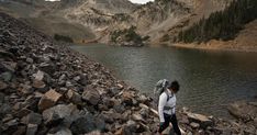 a woman walking on rocks near a lake in the mountains with her backpack over her head