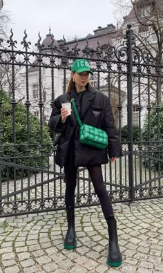 a woman standing in front of a black fence holding a green purse and looking at her cell phone