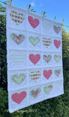 a quilt hanging on a clothes line with hearts in the middle and green bushes behind it