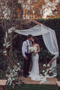 a bride and groom kissing in front of an outdoor wedding ceremony arch with white flowers