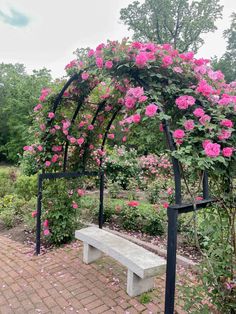 a white bench sitting under a pink rose covered arbor in a park area with flowers all around it