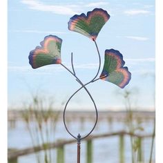 three metal flowers on top of a pole near the water with grass and sky in the background