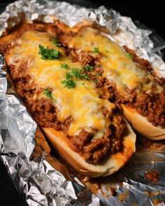 two hot dogs covered in chili and cheese sitting on tin foil with green garnish