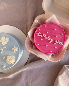 two birthday cakes in plastic containers on a table with pink and white flowers around them