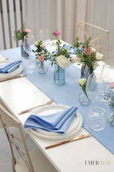 the table is set with blue linens and white plates, gold cutlery, and flowers in glass vases