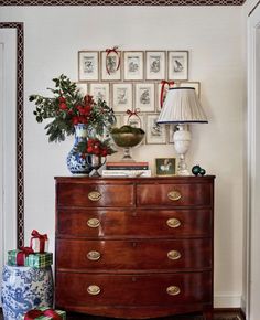 a chest of drawers with christmas presents on the top and below it, in front of a wall with framed pictures