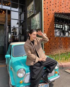 a woman is sitting on the hood of a small car in front of a building