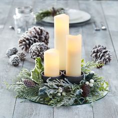 three lit candles are sitting on a table surrounded by pine cones and greenery