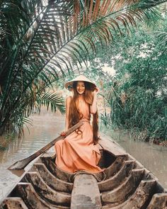 a woman wearing a hat and sitting in a boat on a river with palm trees