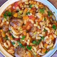 a white bowl filled with pasta and sausage soup on top of a wooden table next to a spoon