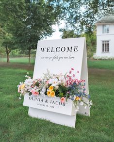 a welcome sign with flowers on it in front of a white house and tree line