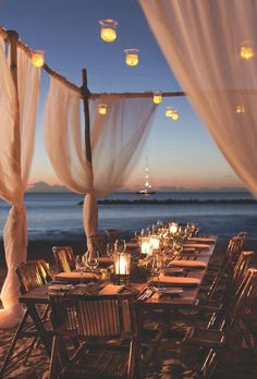 an outdoor dining set up on the beach with candles and lanterns hanging from the ceiling