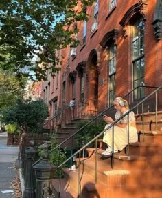 a woman in white dress sitting on steps next to red brick building with trees and bushes