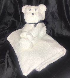 a white teddy bear sitting on top of a black cloth covered bed sheet with rolled up blankets