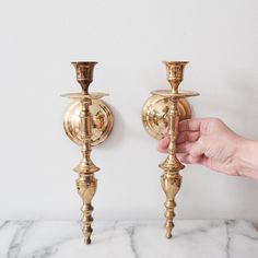 two golden candlesticks on a marble countertop, one being held by a person's hand