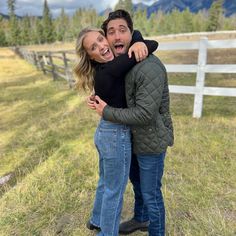 a man and woman hugging in front of a fence with mountains in the background on a cloudy day