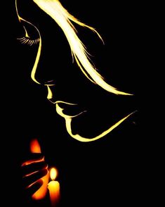 a woman holding a lit candle in her hand and looking down at the ground, with dark background