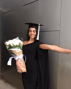 a woman in a graduation cap and gown holding flowers, standing next to a wall