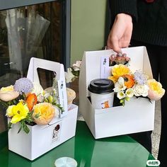 two white boxes with flowers in them sitting on a green countertop next to a person holding a cup