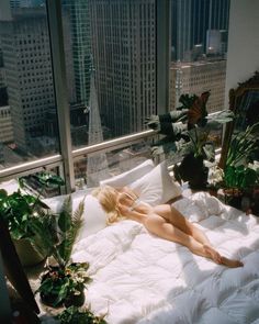 a woman laying on top of a white bed in a bedroom next to tall buildings