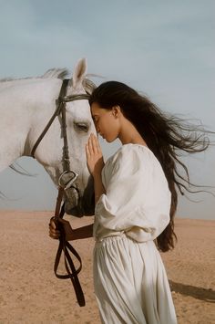 a woman in white dress standing next to a white horse with long hair blowing it's tail