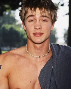 a close up of a shirtless man wearing a chain around his neck with trees in the background