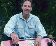 a man holding a pink skateboard in his hands and smiling at the camera with trees behind him