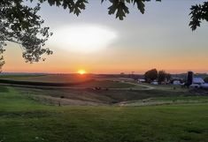 the sun is setting over an open field with farm buildings in the distance and trees to the side