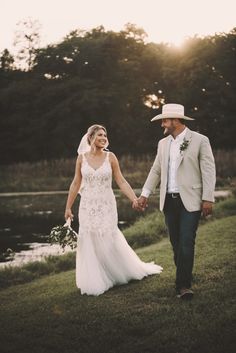 a bride and groom holding hands while walking in the grass near a lake at sunset