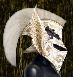 a white and gold mask with feathers on it's head, sitting on a black mannequin