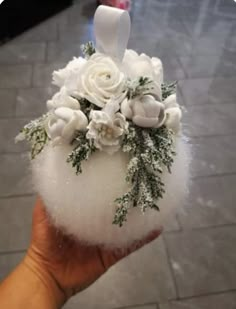 a hand holding a white vase with flowers and greenery on it's side