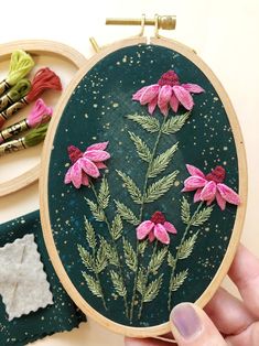 a hand holding a embroidery kit with pink flowers on it and other items in the background