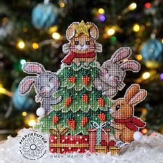 a cross stitch christmas tree ornament with two rabbits sitting on it in front of a decorated christmas tree